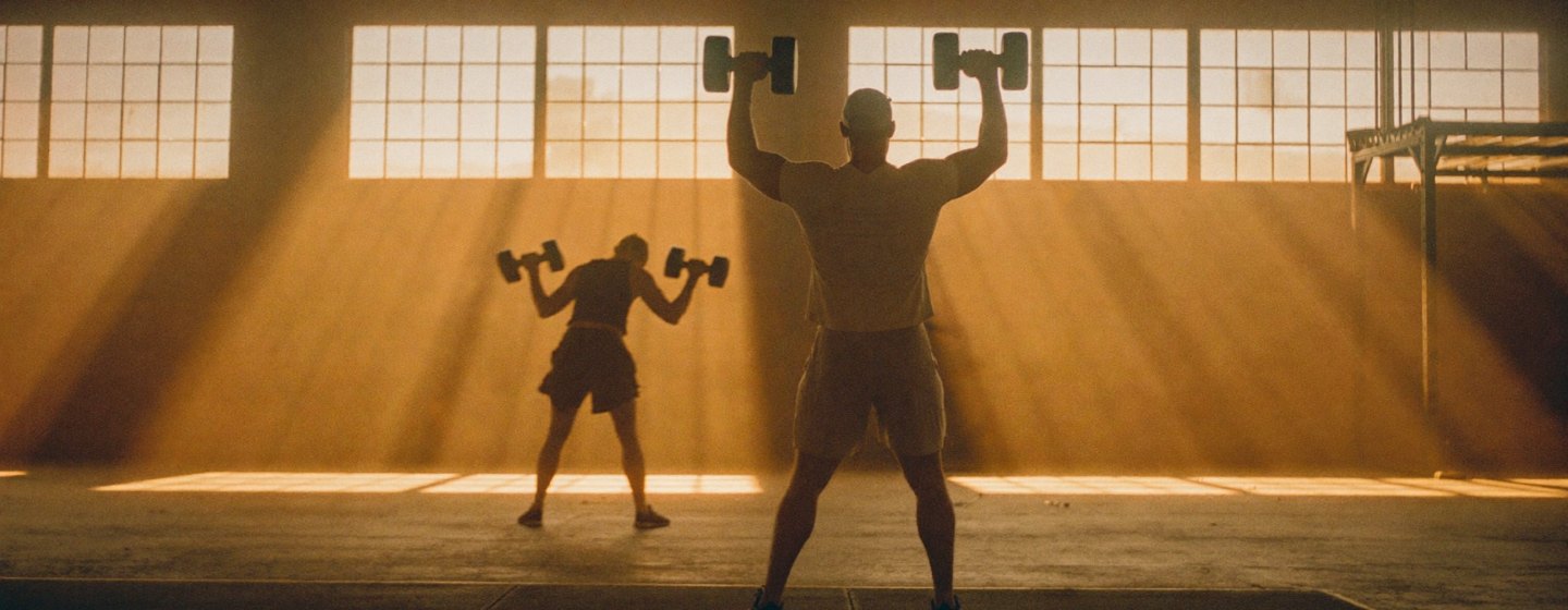 Two people lifting dumbbells in a sunlit warehouse gym